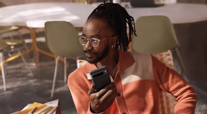 Un homme tient un smartphone noir dans un espace intérieur avec des chaises et une table en arrière-plan.