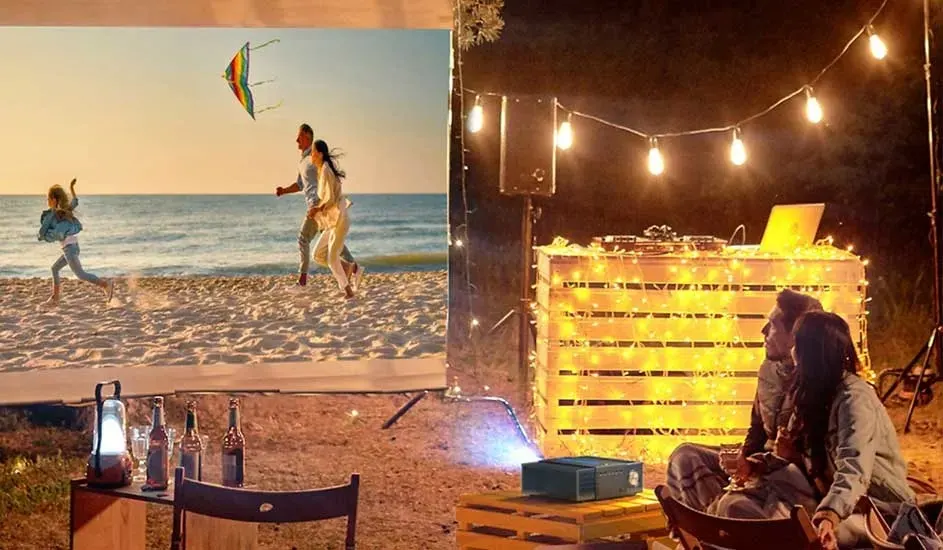 Deux personnes sont assises dehors sur une terrasse illuminée, regardant une projection d’une scène de plage avec un cerf-volant, entourées de bouteilles et de guirlandes lumineuses. Aucune marque n’est visible.