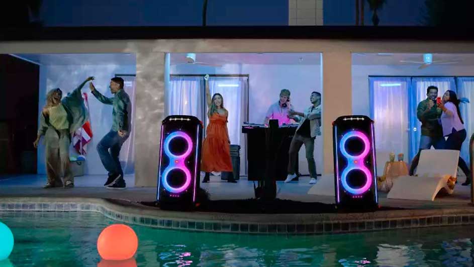 Fête au bord de la piscine avec des haut-parleurs lumineux, des lumières flottantes colorées et des gens qui dansent.