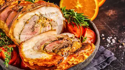 Rôti de porc tranché avec tomates, romarin et orange dans un bol en bois. Fond sombre avec une serviette violette.