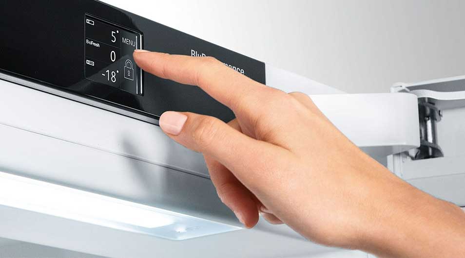 Une main ajuste les paramètres sur le panneau de commande noir d'un réfrigérateur blanc, affichant les options "5", "0" et "-18".