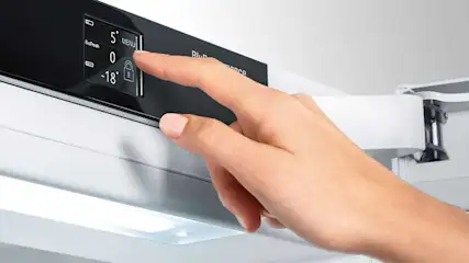 Une main ajuste les paramètres sur le panneau de commande noir d'un réfrigérateur blanc, affichant les options "5", "0" et "-18".