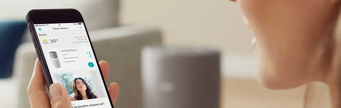 Een vrouw bekijkt op haar smartphone een slimme luchtzuiveraar, terwijl het toestel wazig zichtbaar is in een rustige woonkamer.