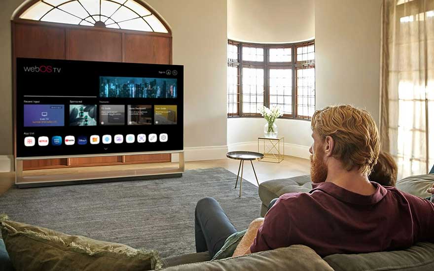 Salon lumineux avec une grande TV noire affichant un menu d'applications. Un canapé gris et une fenêtre cintrée sont visibles.