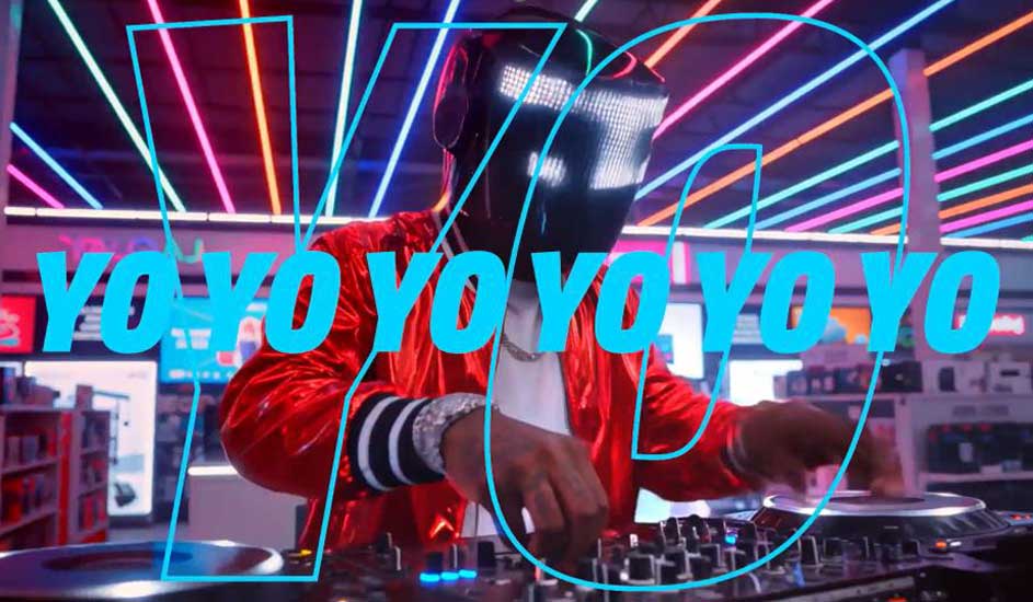 Un DJ avec un masque lumineux mixe dans un magasin, entouré de néons colorés et le mot « YO YO YO YO YO YO » affiché.
