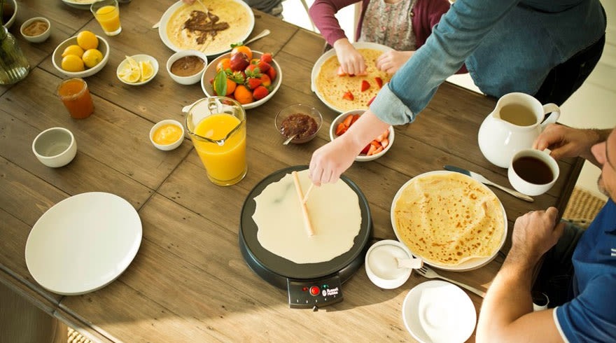 Table en bois avec crêpes, fruits, jus, et machine à crêpes. Assiettes blanches, bols avec garnitures variées. Préparation de crêpes en cours.