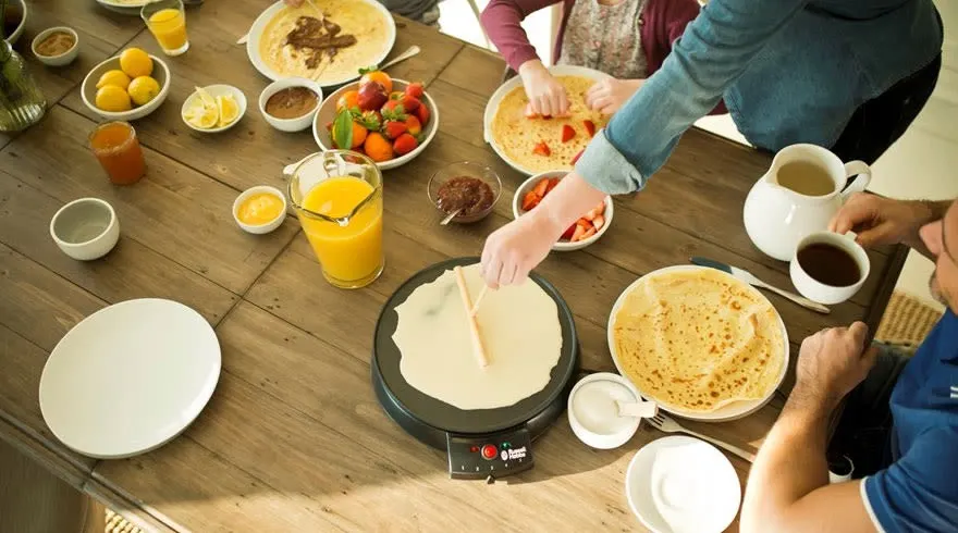 Table en bois avec crêpes, fruits, jus, et machine à crêpes. Assiettes blanches, bols avec garnitures variées. Préparation de crêpes en cours.