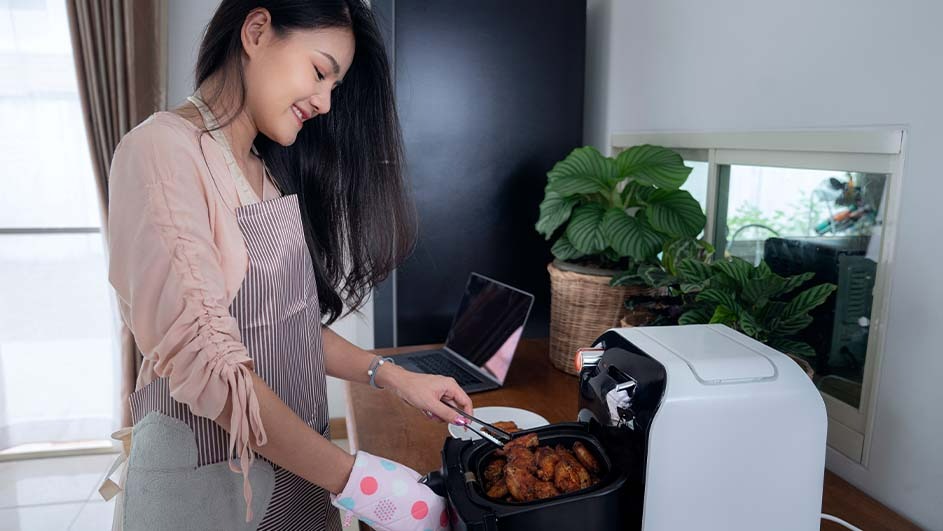 Een witte airfryer staat op een aanrecht met eten erin, met een laptop, planten en een raam op de achtergrond.