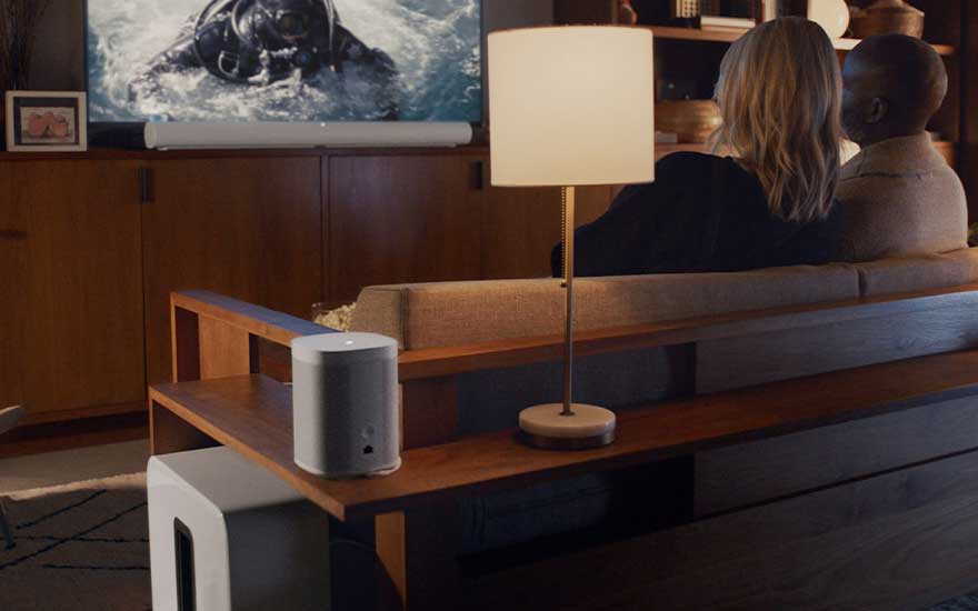 Woonkamer met tv, soundbar, houten kast, lamp, witte speaker en een bank. Een blauw tapijt is deels zichtbaar.