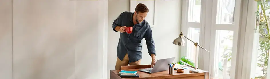 Un homme à un bureau en bois avec ordinateur, lampe en métal et tasse rouge, près d'une fenêtre lumineuse.