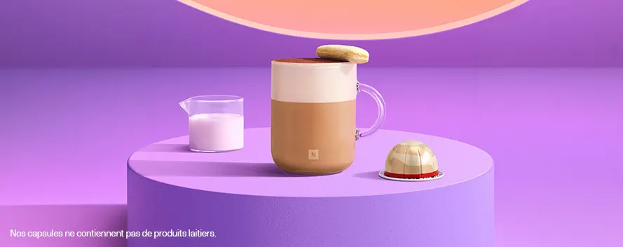 Une boisson au café crémeuse et mousseuse dans une tasse en verre sur un socle violet, à côté d’un petit pichet de lait, d’un biscuit et d’une capsule dorée. Une note en bas indique que les capsules ne contiennent pas d’ingrédients laitiers.
