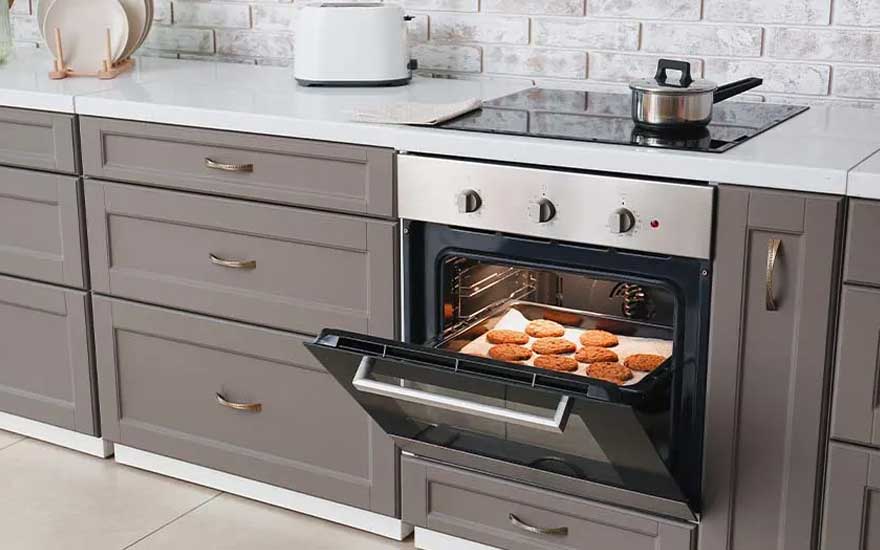 Open oven met koekjes op een bakplaat, grijze kasten en een witte aanrecht met een broodrooster en kookpot.
