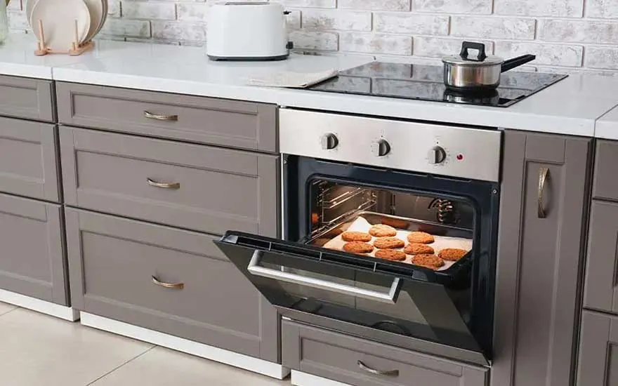 Open oven met koekjes op een bakplaat, grijze kasten en een witte aanrecht met een broodrooster en kookpot.