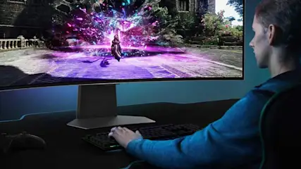 Een gebogen monitor toont een videospel met paarse effecten. Een toetsenbord en controller liggen op het bureau eronder.