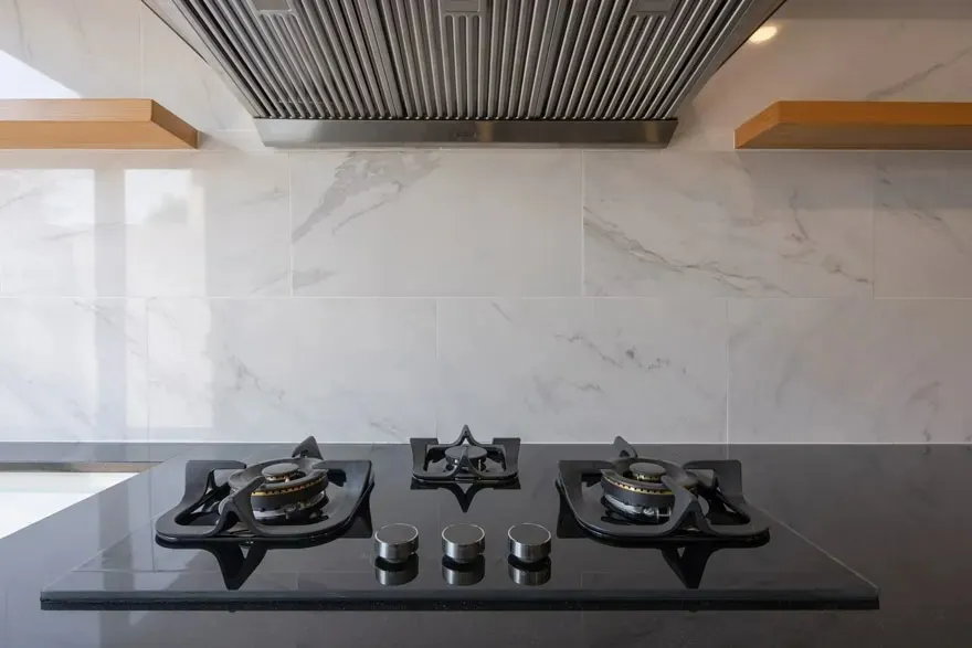 Vue d'une cuisine avec une plaque de cuisson noire à trois brûleurs sous une hotte en acier inoxydable.