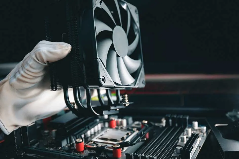 Een hand met een witte handschoen houdt een zwarte CPU-koeler boven een moederbord, met zichtbare rode en zilverkleurige componenten.