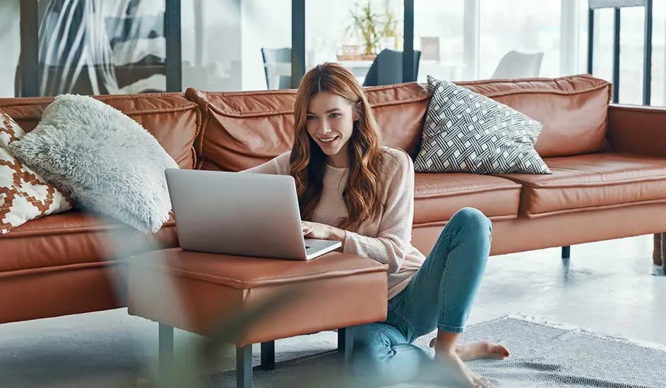 Woman sits on floor in front of brown leather sofa, happily using laptop on ottoman in bright modern living room.