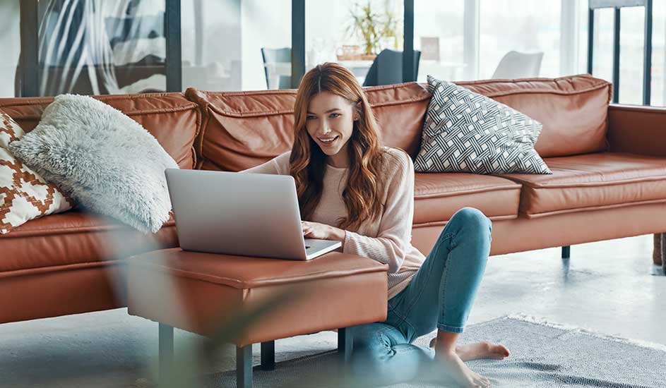 Vrouw zit op de grond voor bruine leren bank, werkt lachend op laptop op hocker in lichte, moderne woonkamer.