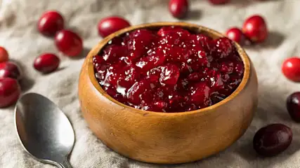 Een houten kom gevuld met rode cranberrysaus staat op een stoffen ondergrond met een zilveren lepel en losse cranberries.