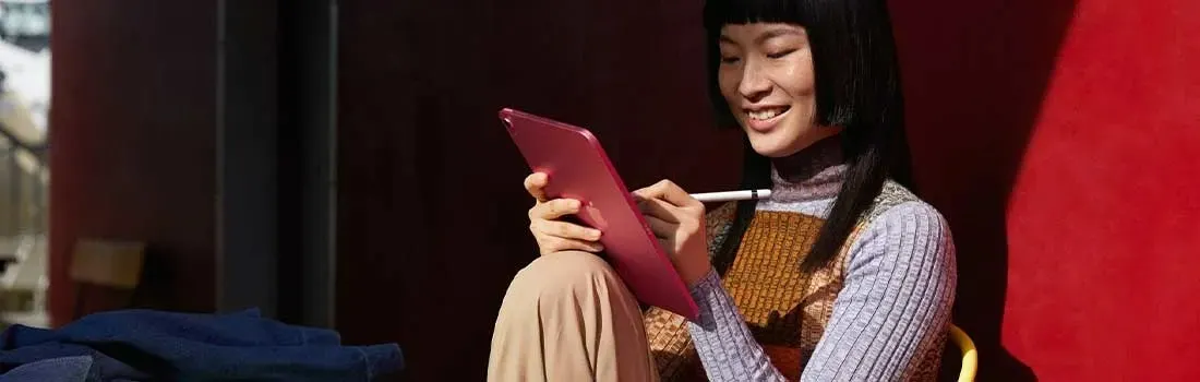 A person using a pink iPad with a stylus, seated against a red wall.