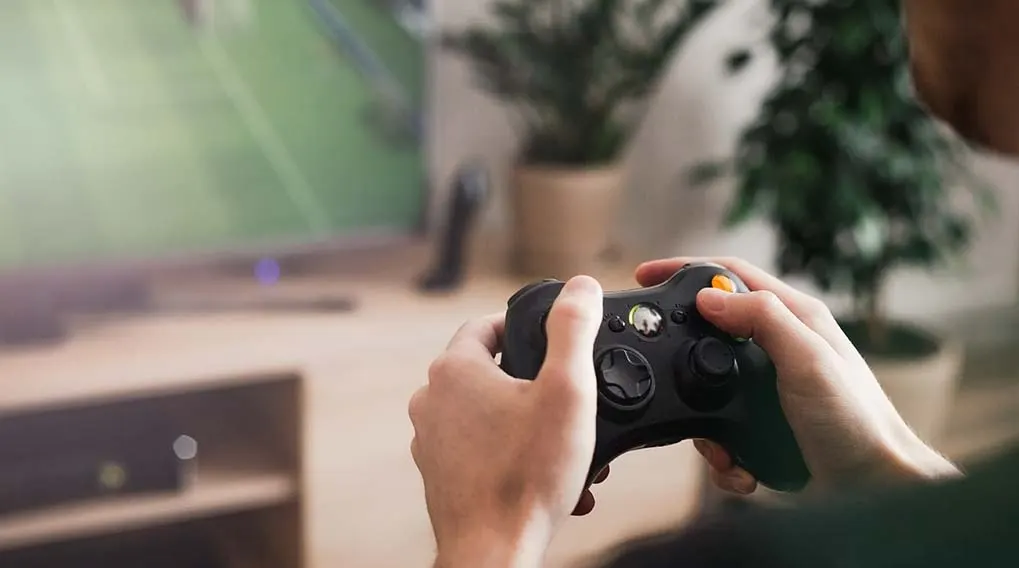 Une personne tient une manette de jeu noire. Un écran de télévision est visible en arrière-plan, ainsi que des meubles en bois et une plante.