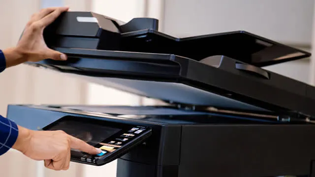 Une personne utilise une imprimante noire multifonction, levant le couvercle et appuyant sur l'écran de contrôle.