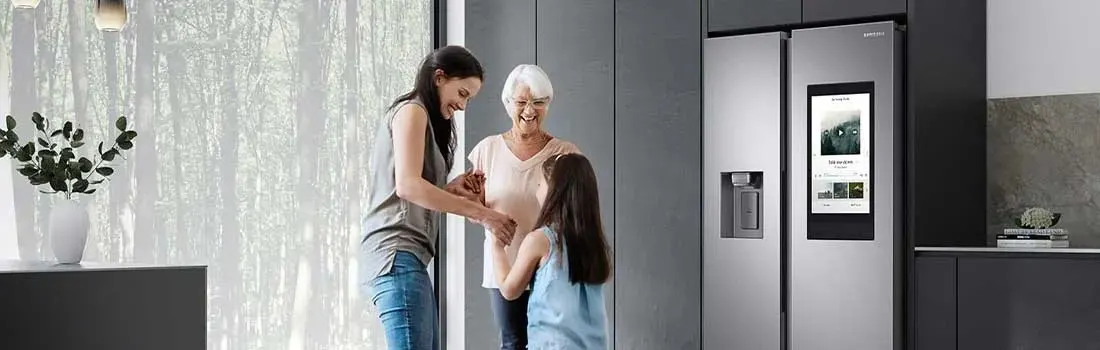 A kitchen features a stainless steel Samsung refrigerator with a screen, next to grey cabinets. A vase with greenery sits on a countertop.