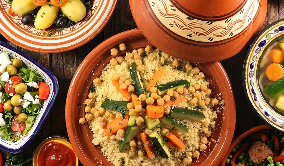 Een bovenaanzicht van verschillende gerechten, waaronder couscous in een tajine, salade en aardappelen op een houten tafel.