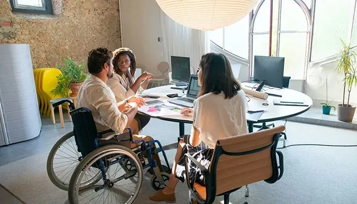 Inclusive meeting room with three colleagues around a table. One person in a wheelchair participating in the discussion in a bright, modern space.