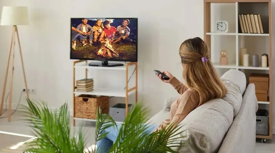 A woman sits on a light sofa watching a TV showing a group of friends around a campfire. No brand is visible on the television.