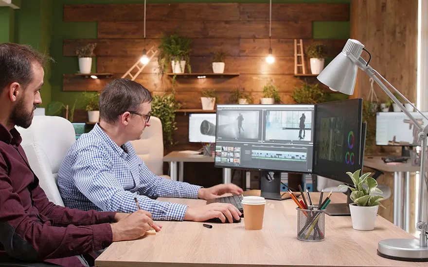 Deux personnes sont assises à un bureau devant des écrans d'ordinateur montrant des logiciels de montage vidéo, dans un bureau décoré de plantes.