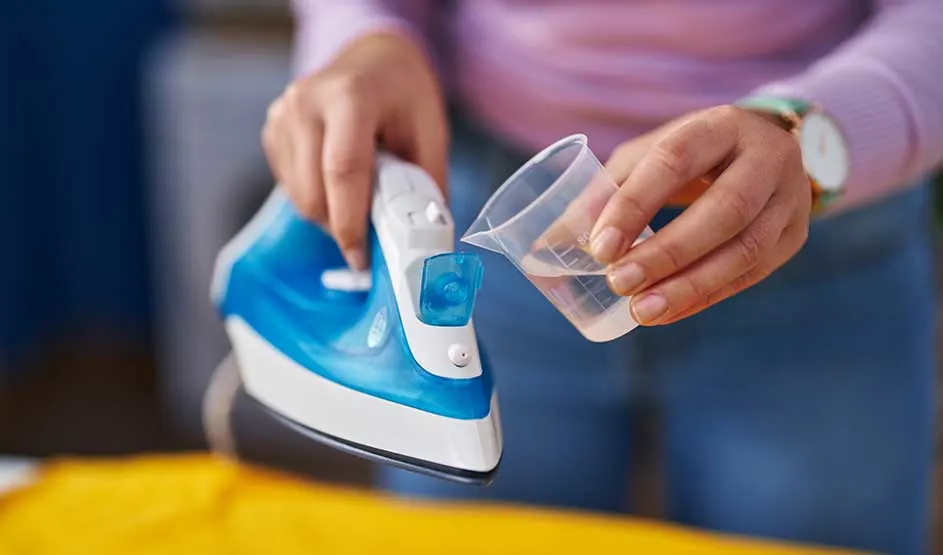 Quelqu’un remplit un fer à vapeur bleu et blanc avec de l’eau versée d’un petit gobelet doseur, un vêtement jaune flou au premier plan.