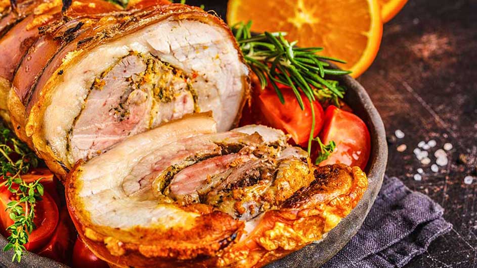 Een close-up van een gebraden rollade op een schaal met tomaten, rozemarijn en sinaasappelschijfjes.