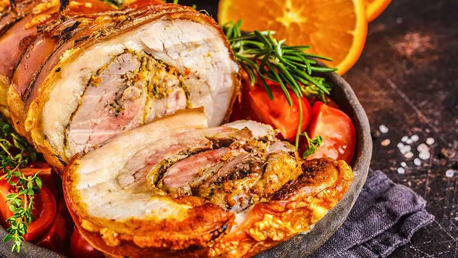 Een close-up van een gebraden rollade op een schaal met tomaten, rozemarijn en sinaasappelschijfjes.