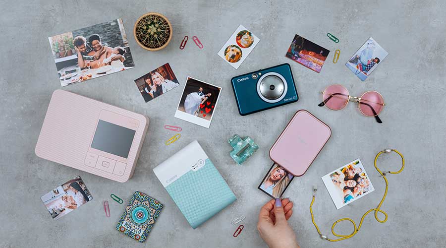 Flatlay met mini fotoprinters, een compacte camera en uitgeprinte snapshots; hand trekt foto uit een roze printer.