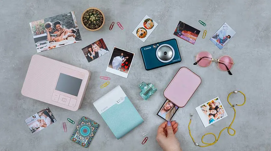 Flatlay met mini fotoprinters, een compacte camera en uitgeprinte snapshots; hand trekt foto uit een roze printer.