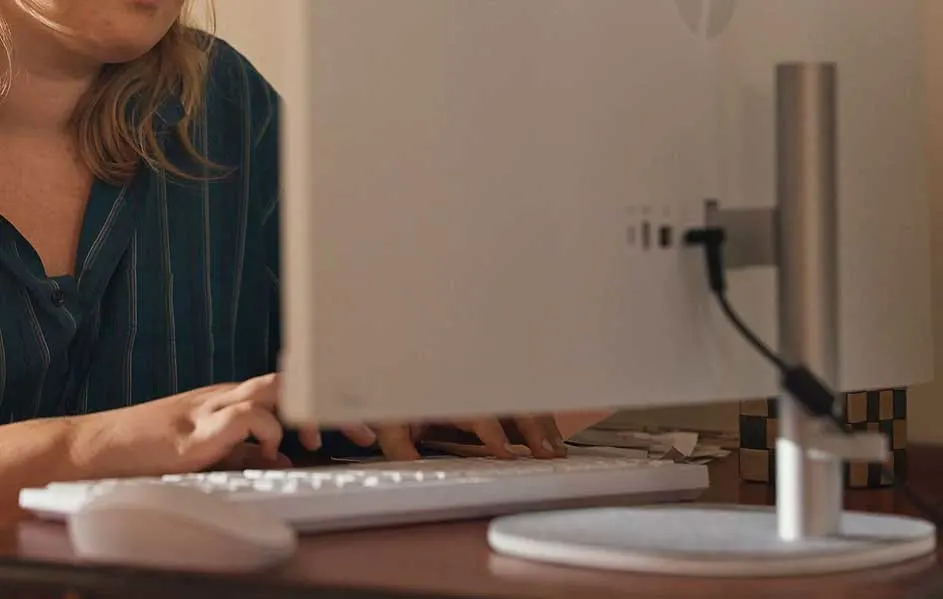 Een wit toetsenbord en een computermuis staan op een houten bureau voor een computerscherm. Kabels zijn aangesloten op de achterkant van de monitor.