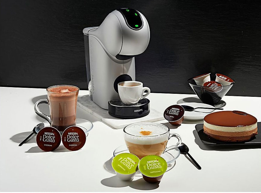 Machine à café grise avec tasses de boissons et capsules Dolce Gusto sur table blanche. Gâteau au chocolat à droite.