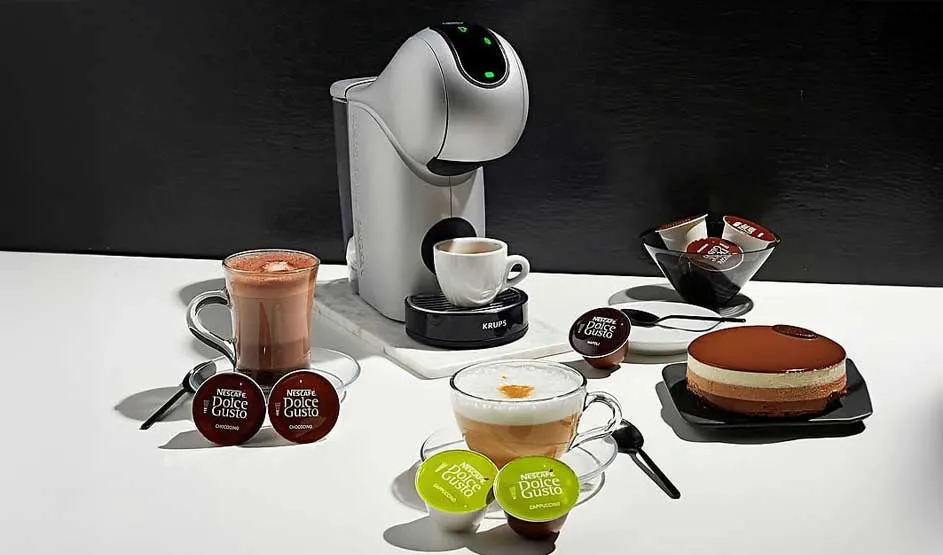 Coffee machine with coffee cups, pods, cake, and spoons on a white table against a black background.