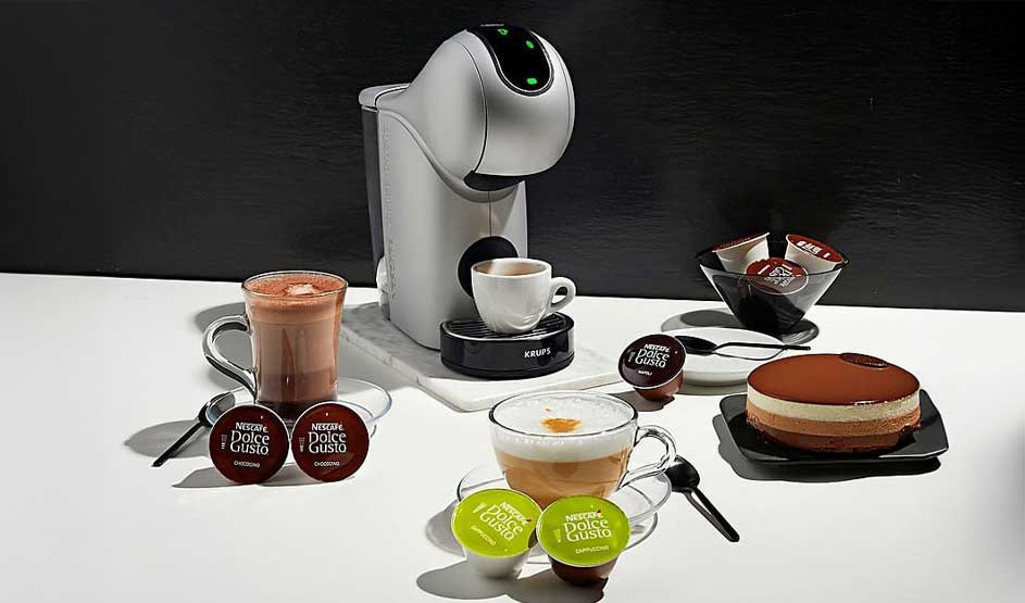 Machine à café grise avec tasses de boissons et capsules Dolce Gusto sur table blanche. Gâteau au chocolat à droite.
