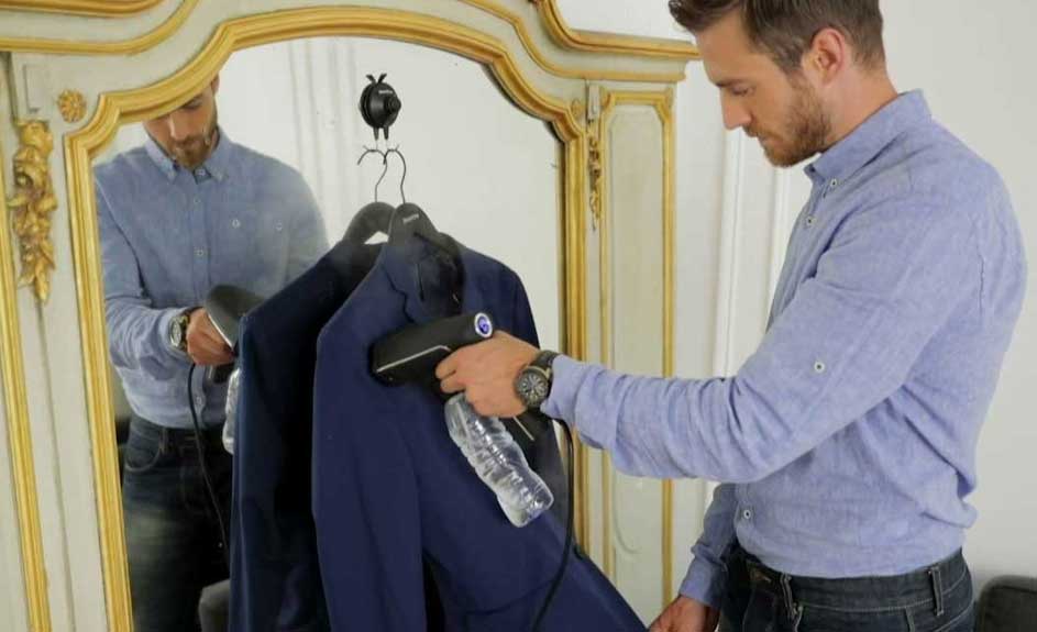 Un homme défroisse une veste bleue suspendue avec un défroisseur noir près d'un miroir orné. Il porte une chemise bleue et un jean.