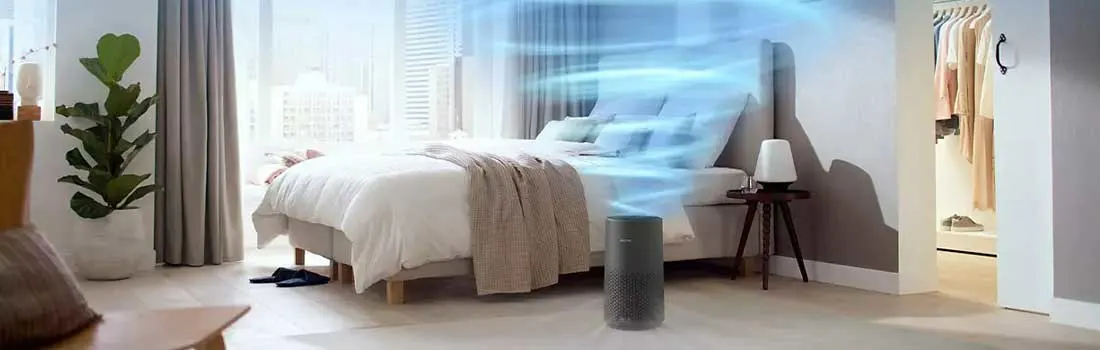 Chambre lumineuse avec lit blanc, purificateur d'air gris, table de chevet en bois, et plante verte près de la fenêtre.
