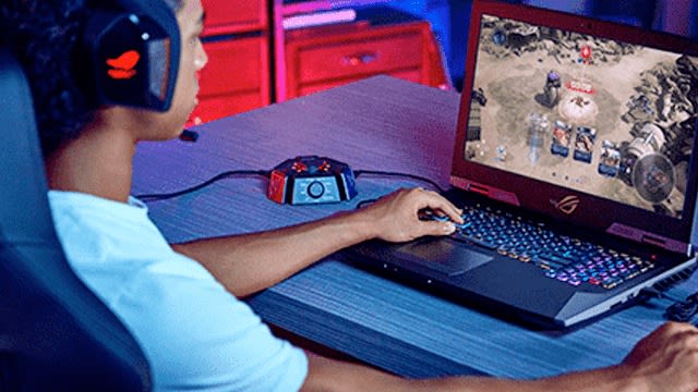 Een persoon met een headset speelt een strategiespel op een laptop op een blauwe houten tafel.