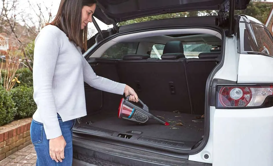 Une femme nettoie le coffre d’une voiture blanche avec un aspirateur à main ; le hayon est ouvert pendant le nettoyage.