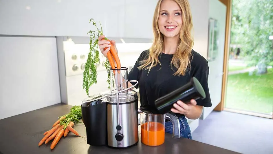 Une centrifugeuse en inox sur un comptoir, avec des carottes et du jus d'orange.