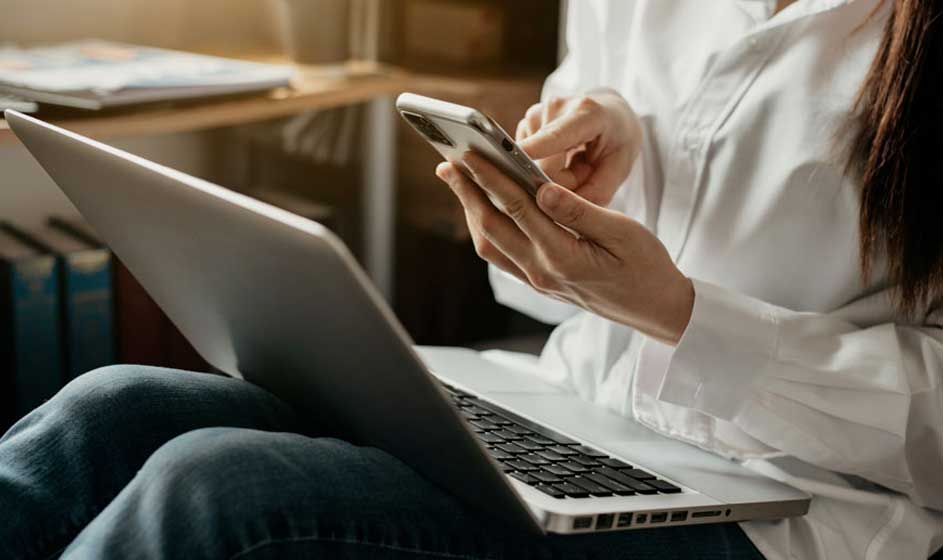 Een laptop rust op blauwe jeans, terwijl iemand een zilverkleurige smartphone bedient. Op de achtergrond staat een houten bureau.