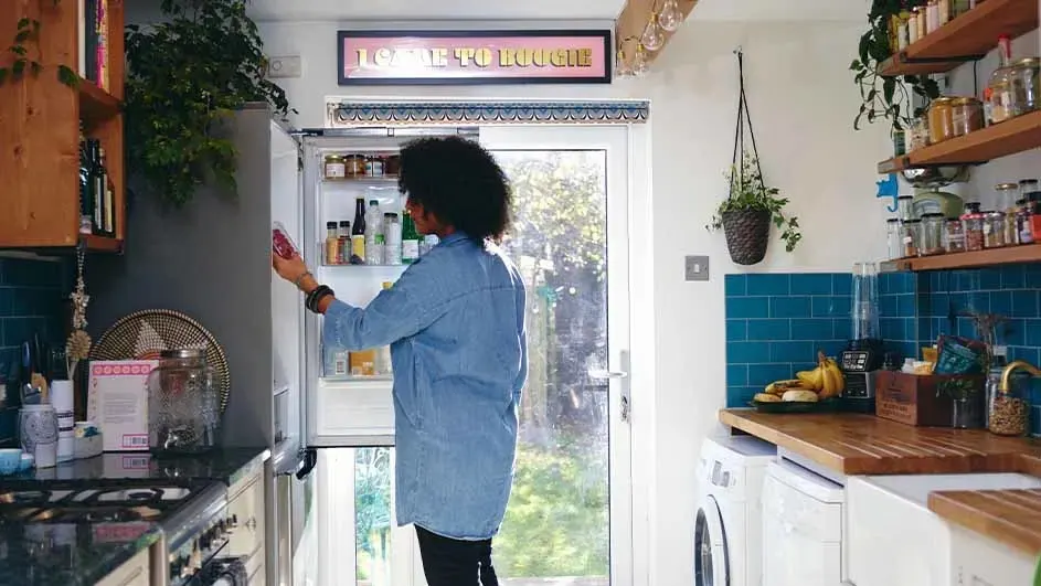 Keuken met houten rekken vol potten en flessen. Een koelkast staat open, gevuld met diverse producten. Een raam toont een groene tuin.