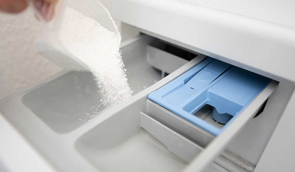 Une personne verse de la lessive en poudre dans un tiroir de machine à laver blanc avec un compartiment bleu.