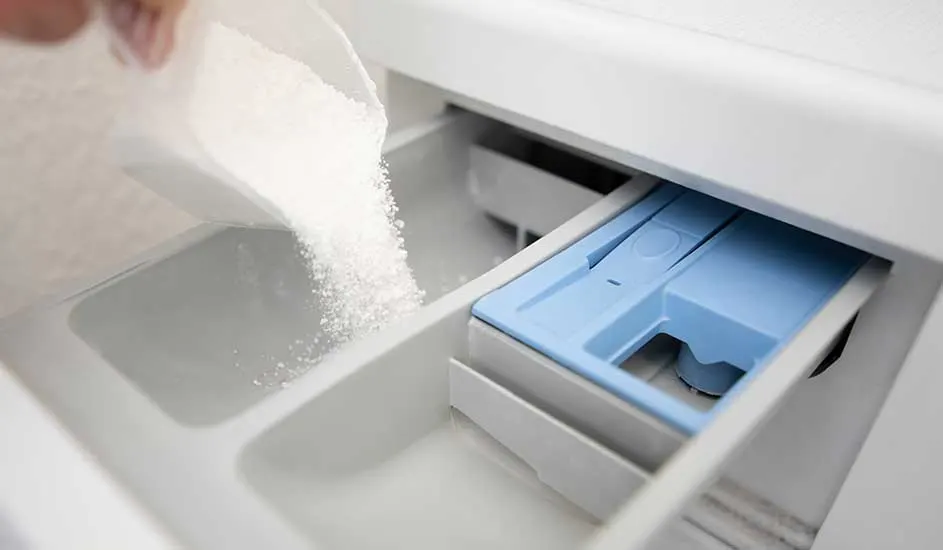 Wasmiddel wordt in een wasmachinebak gegoten. De bak heeft een blauw compartiment.