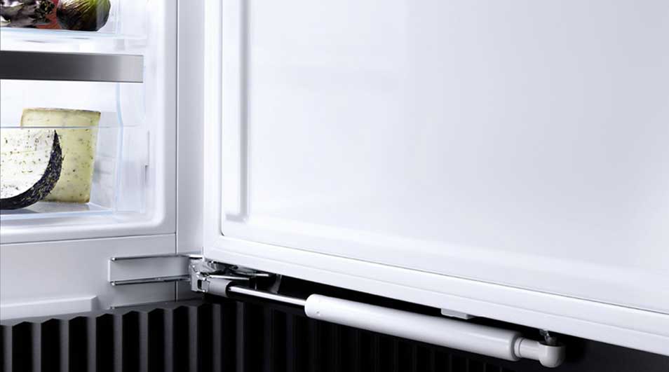 Un frigo blanc ouvert montre du fromage et des fruits sur une étagère. Un mécanisme de fermeture est visible en bas.